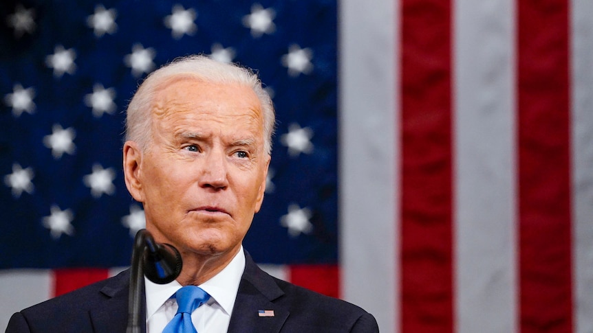 Joe Biden speaking into a microphone in front of an American flag