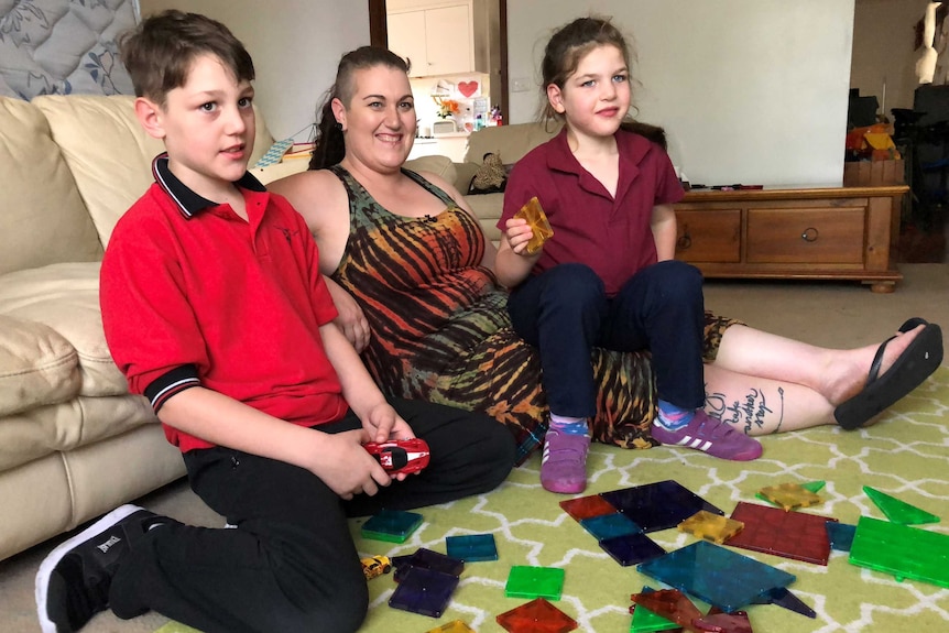 Mum Sarah Chapman smiles as she sits on the ground between son Deegan, 8, and daughter Ayla, 6, next to some toys on the floor.