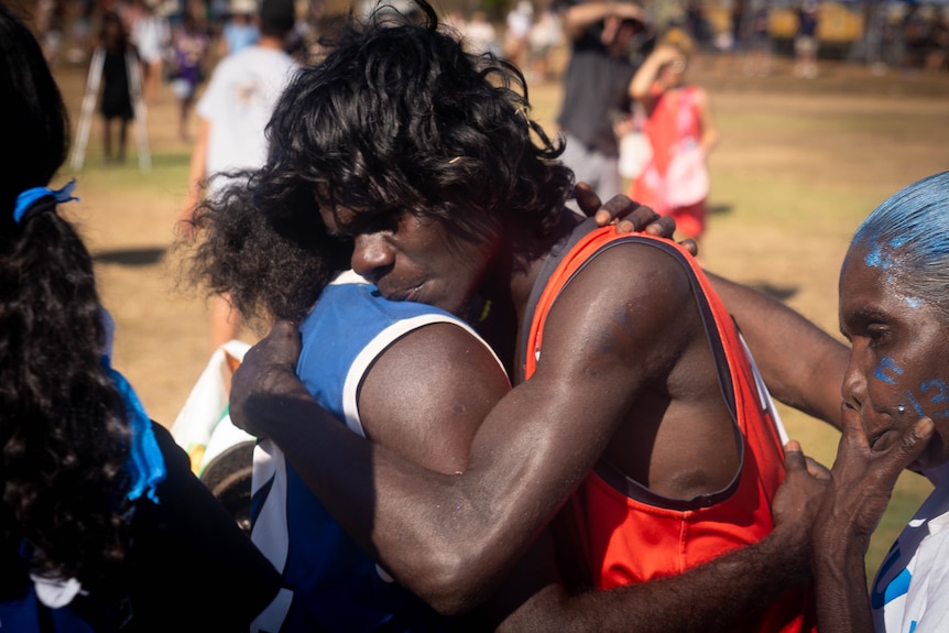 Two young Indigenous men embrace, each wearing different jerseys.