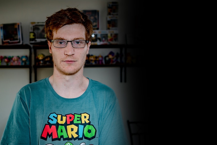 A young man in a room, with a shelf of toys in the background.