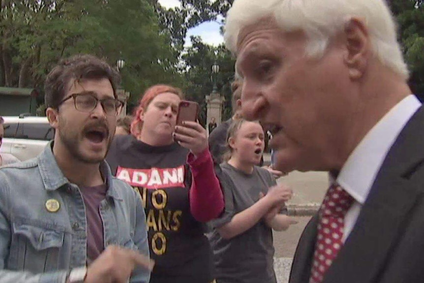A man with a group of people behind him yelling at Bob Katter