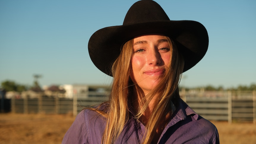 a young woman stands smiling in a purple shirt and a cowgirl hat in the outback