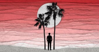 A man looks out to sea behind palm trees lit by a red sky