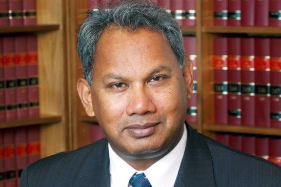 Justice Rangiah stands in front of bookshelves with leather-bound red law books on them.