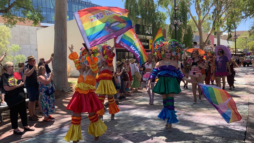Three performers dressed in colourful 'drag' wave rainbow flags and walk down a mall while people look on smiling.