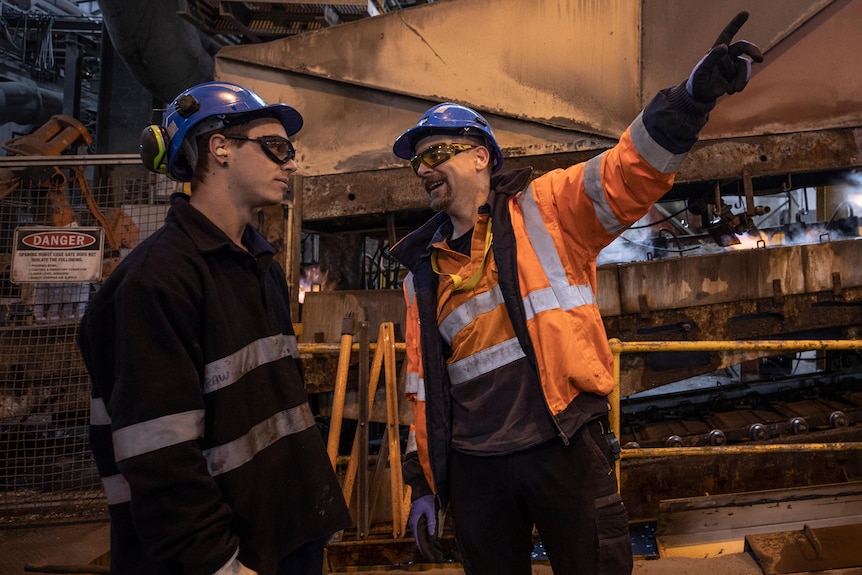 Two men in hard hats and work gear stand in front of machinery.