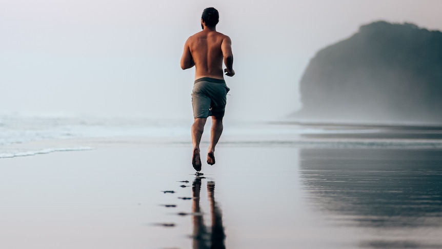 A back view of a shirtless barefoot man running along an empty beach