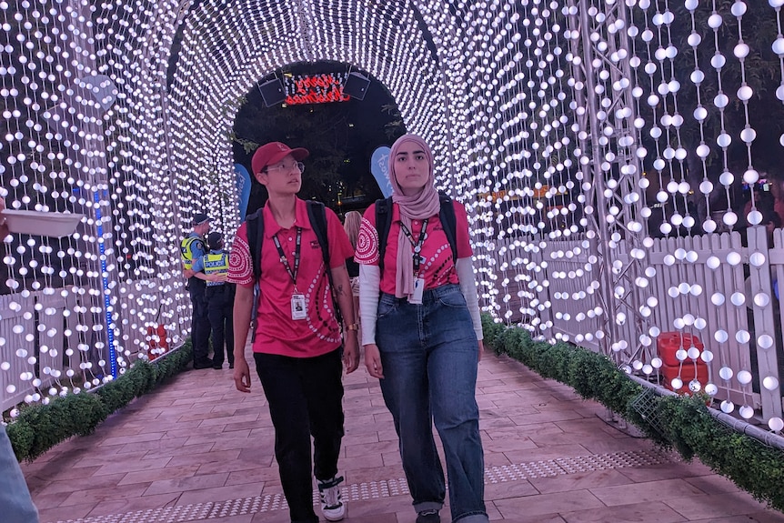 Two women in pink shirts walk through an archway-tunnel of lights in Northbridge 