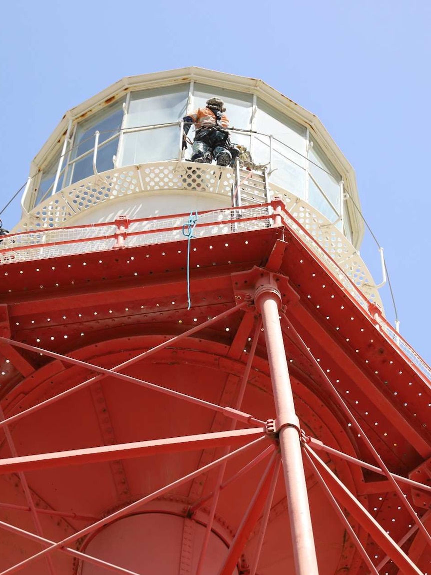 A red and white metal structure towers in a blue sky. At the top of the structure a man is cleaning windows wearing a harness.