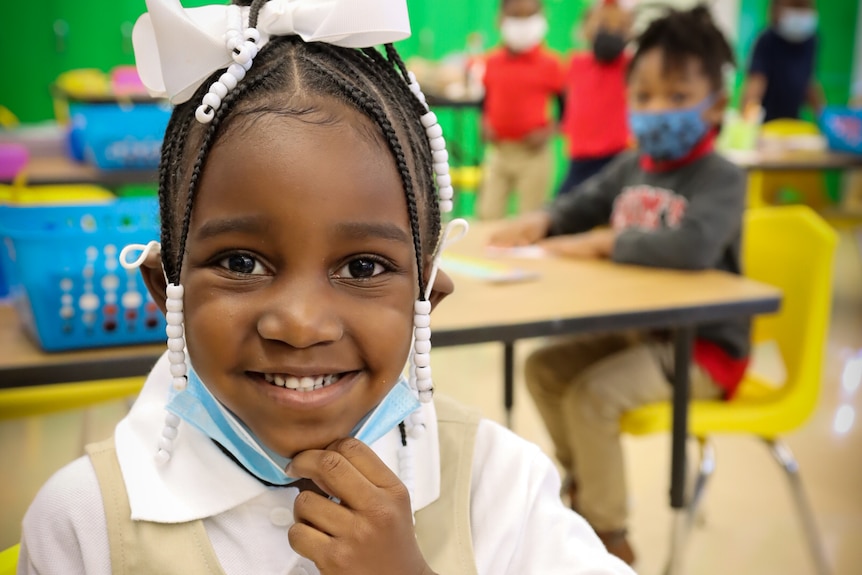 A little girl pulls her face mask down in a classroom and grins 