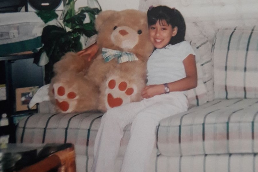 A smiling tween girl dressed in white sits on a sofa with a large teddy bear