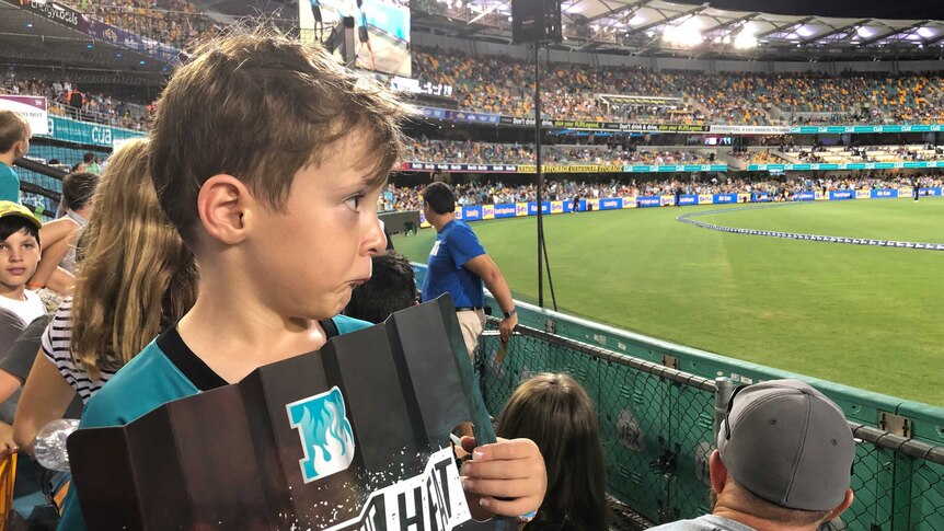 A young boy in Brisbane Heat gear, holding a sign reading "BRING THE HEAT" looks sad after a Gabba BBL game was called off.