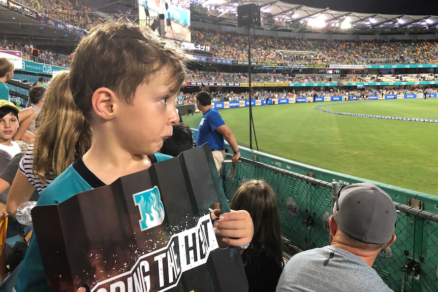 A young boy in Brisbane Heat gear, holding a sign reading "BRING THE HEAT" looks sad after a Gabba BBL game was called off.