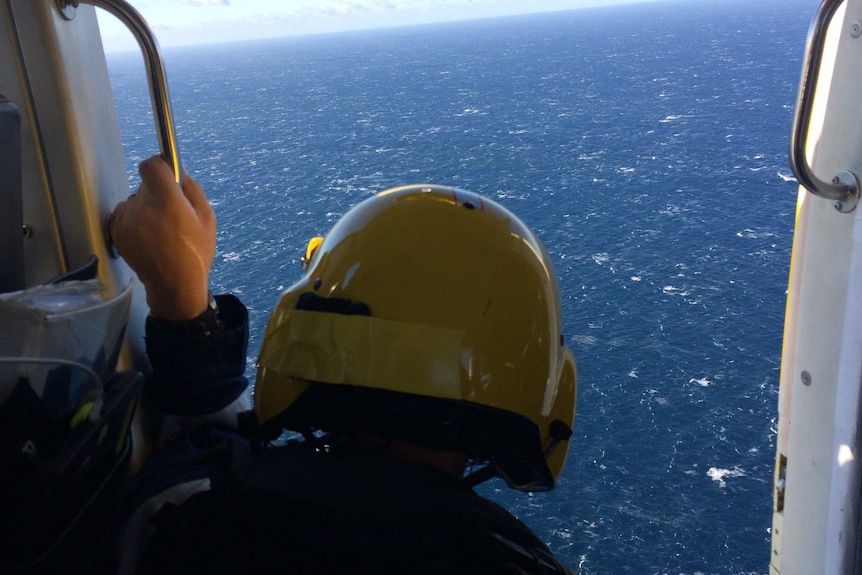 A man in a helmet sits on the edge of a helicopter looking down to sea