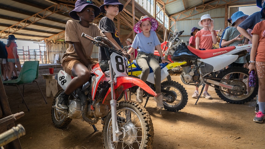 A group of kids sit on motorbikes in a shearing shed
