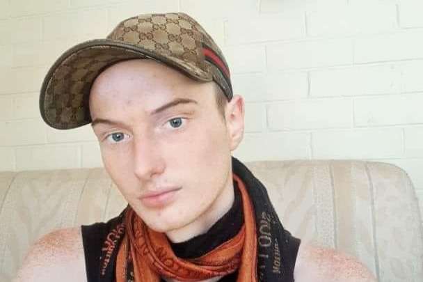 Young Caucasian man taking a self portrait on a couch wearing a brown cap, a black singlet and an orange scarf. 