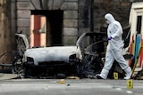 A forensic specialist in protective gear works the scene of a car bombing, placing markers around wreckage strewn across a road.