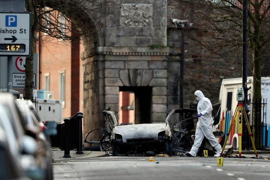 A forensic specialist in protective gear works the scene of a car bombing, placing markers around wreckage strewn across a road.