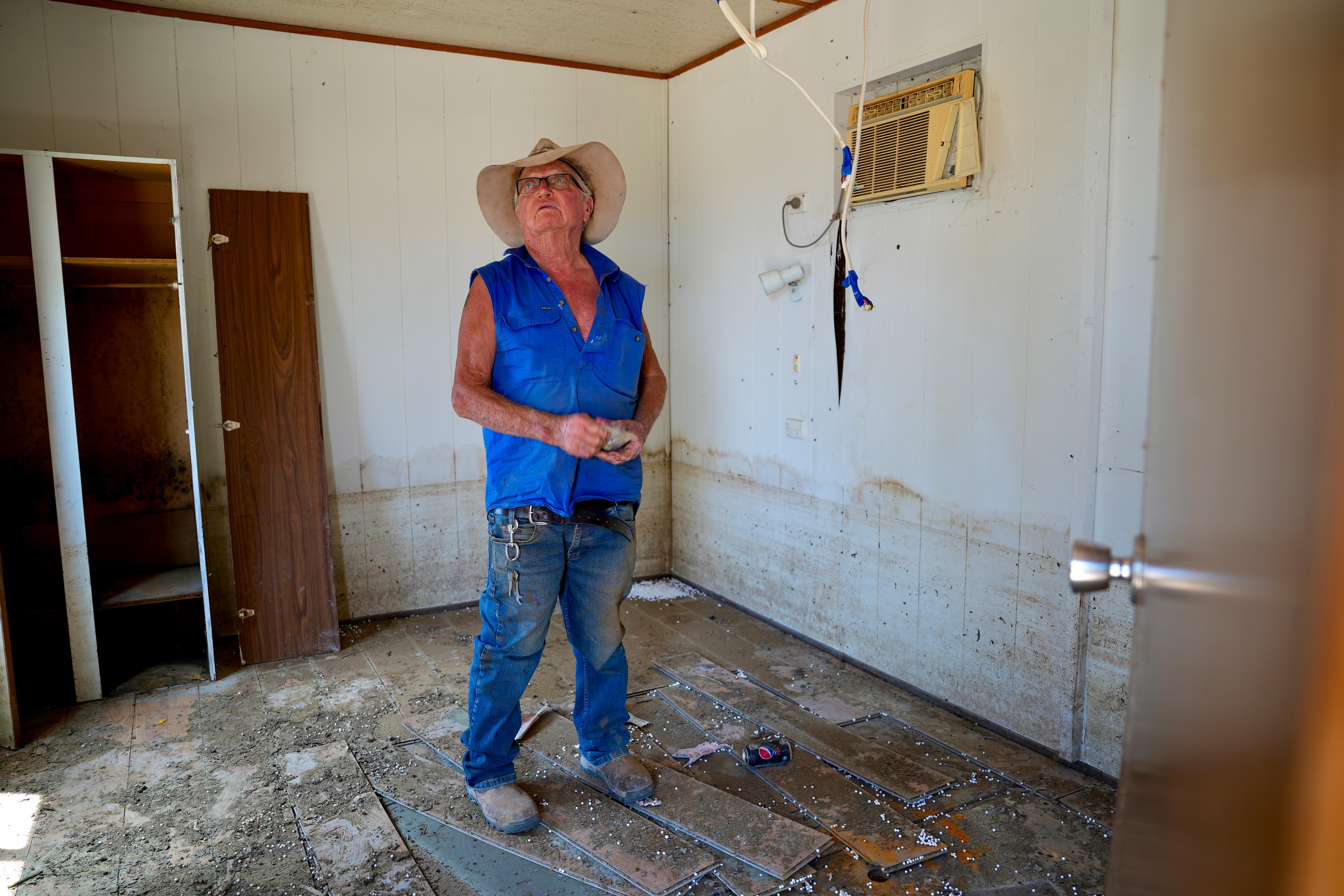 An old white man with a blue sleeveless top, a hat and glasses stands in a flood damaged room near Menindee. 