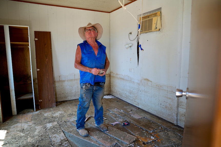 An old white man with a blue sleeveless top, a hat and glasses stands in a flood damaged room near Menindee. 