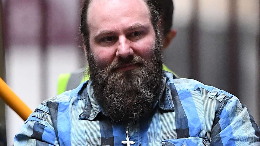 A close-up photo of Phillip Galea, wearing a checked blue shirt and a visible cross necklace, being escorted into court.