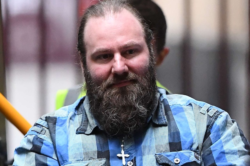 A close-up photo of Phillip Galea, wearing a checked blue shirt and a visible cross necklace, being escorted into court.