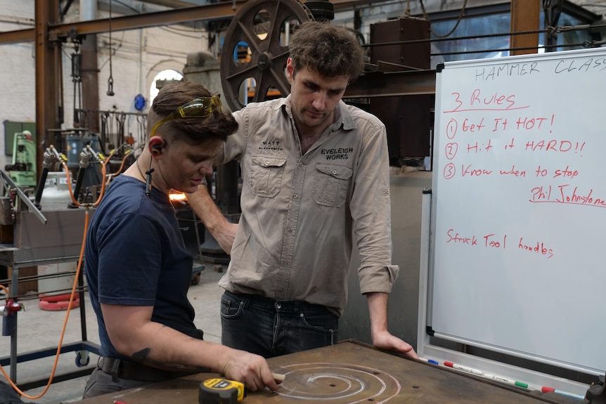 A man and a woman look down at a chalk drawing of a spiral on a table inside an industrial workshop
