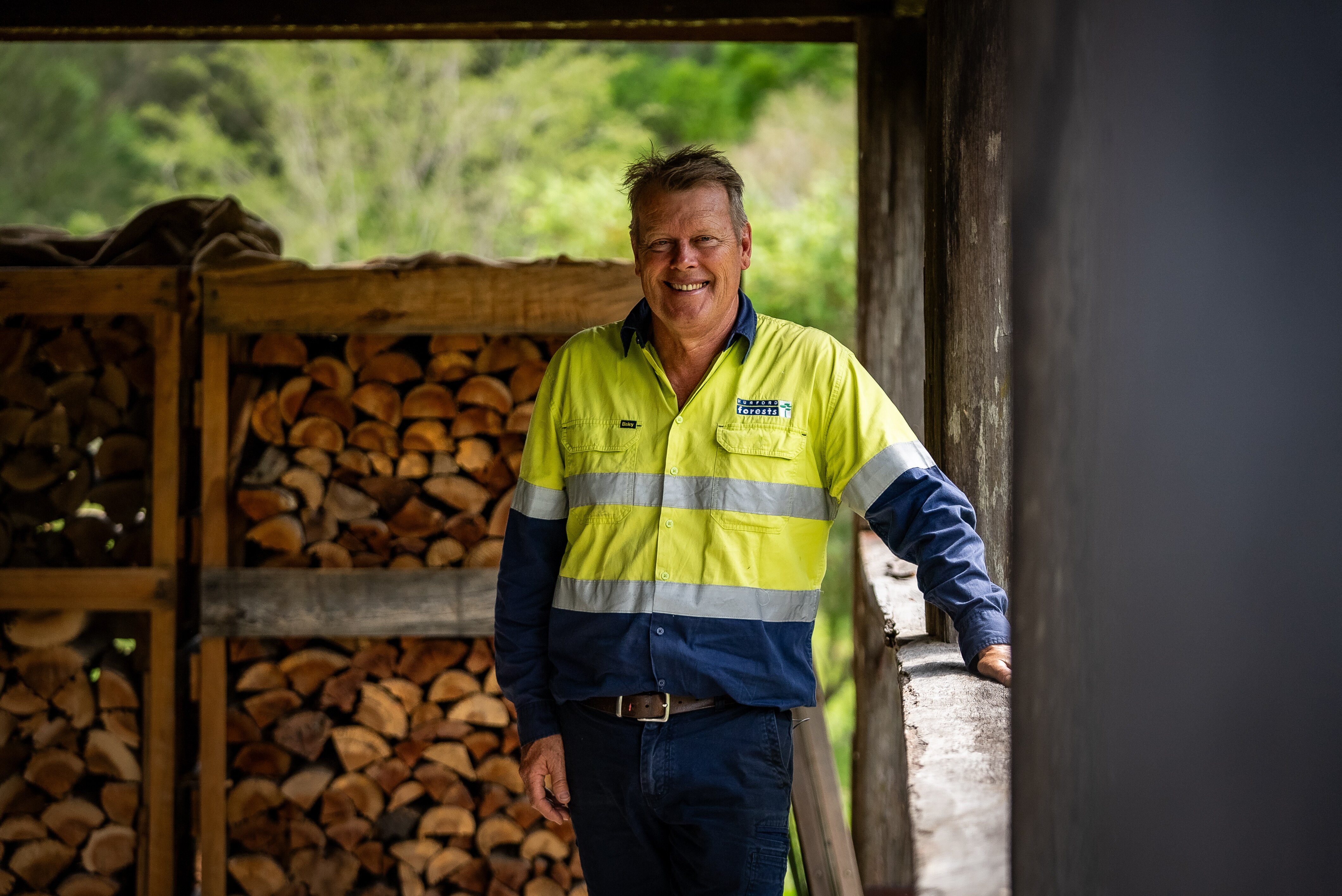 Um homem com uma camiseta de alta visibilidade sorrindo e apoiado em uma grade de madeira, com uma coleção de lenha empilhada atrás dele.