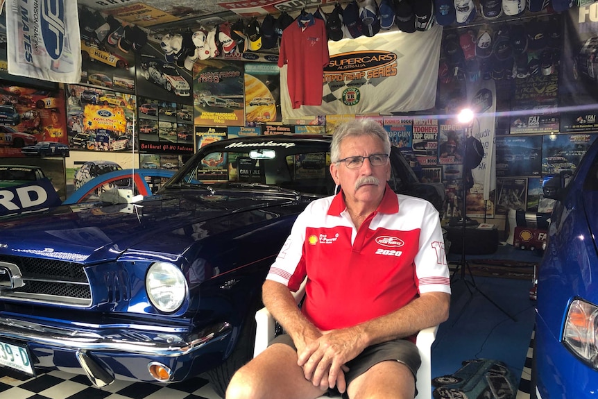 A man sits in a room lined with posters, hats and memorabilia of V8 supercars.