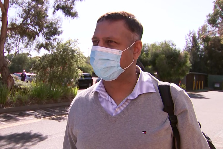 A man in a light grey jumper over a blue striped shirt, wearing a face mask