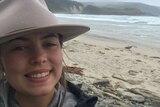 selfie of a young, smiling woman wearing hat with the beach behind her