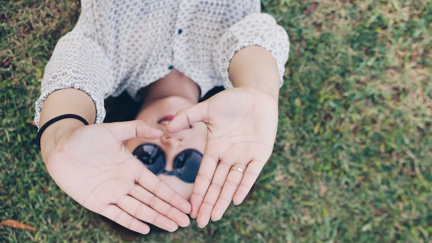 A woman lies in the grass, holding her hands towards the camera in a way that frames her smiling face.