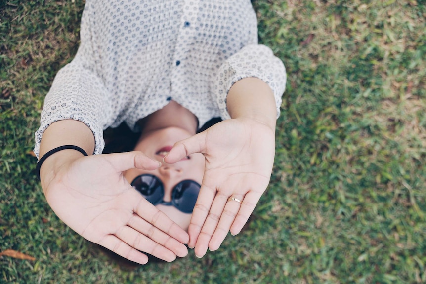 A woman lies in the grass, holding her hands towards the camera in a way that frames her smiling face.