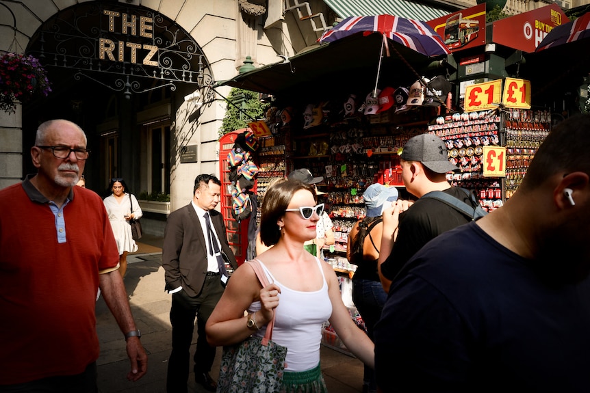 A woman in white cat-eye sunglasses and a singlet walks through a busy London street 
