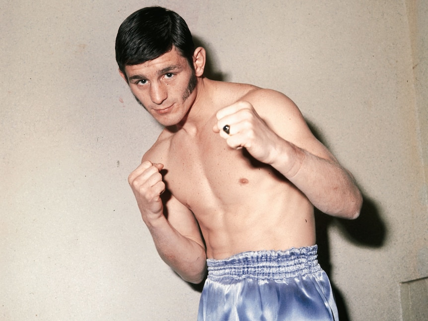 Un boxeador posa para una foto con el torso desnudo, en calzoncillos, con los puños cerrados, frente a la cámara.