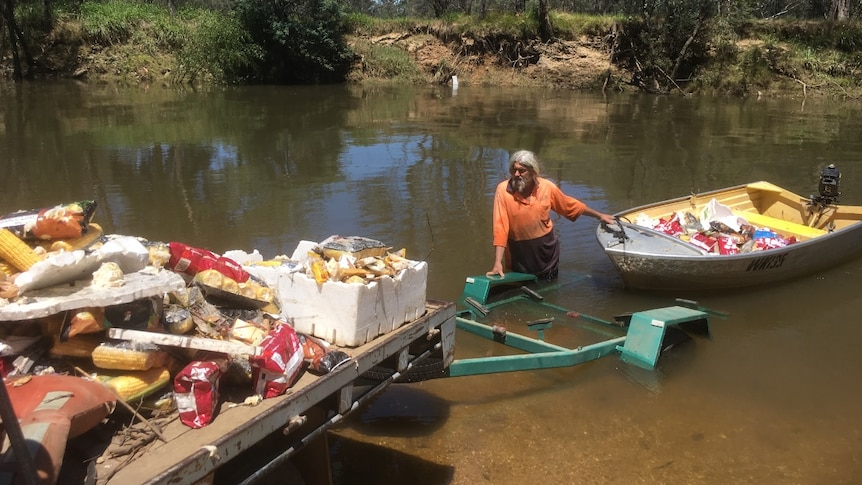 A man stands in a river with a boat filled with rubbish and a trailer stands on nearby land filled with debris.