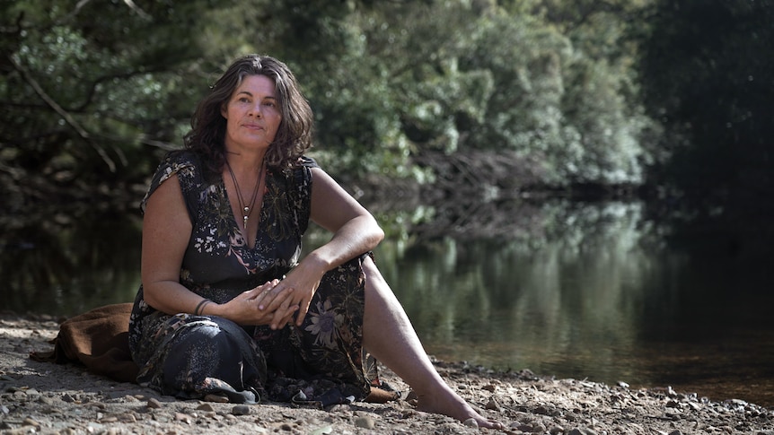 Gina Chick sitting on the ground, water and trees behind her. 