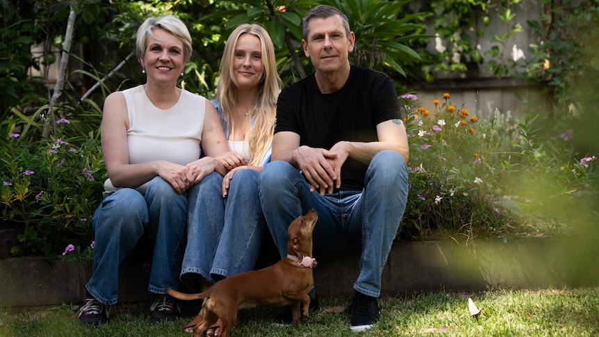 Man woman and 23-year-old daughter sit outside on the grass. their small brown dog at their feet