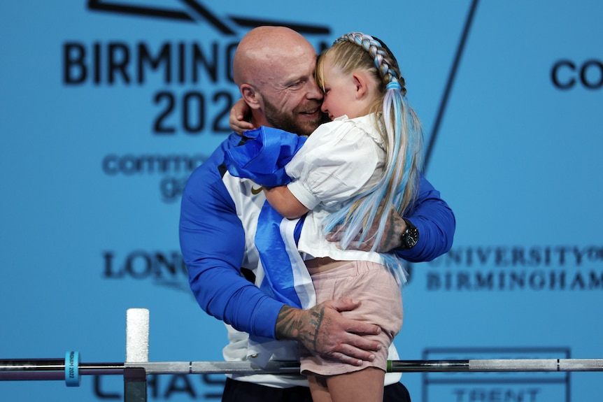 A man wearing a blue and white jacket hugs his daughter.  She has long, blonde hair pulled back in Dutch braids and blue ribbons