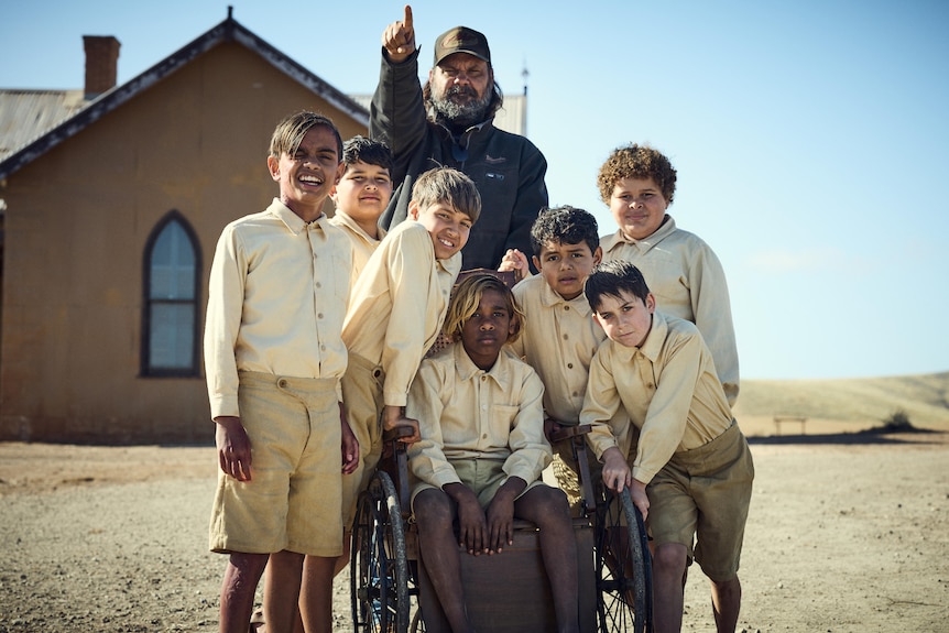 A group of children, some Aboriginal stand around a boy in a wheelchair. A church is seen behind them.