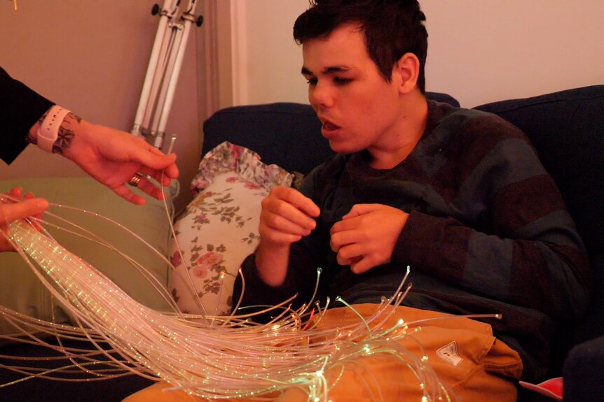 Luke sitting with fibre optic lights across his lap, two hands reaching out beside him.