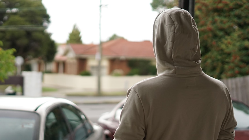 A man in a beige hoodie looks out on a suburban street, with his back to the camera and his face not visible.
