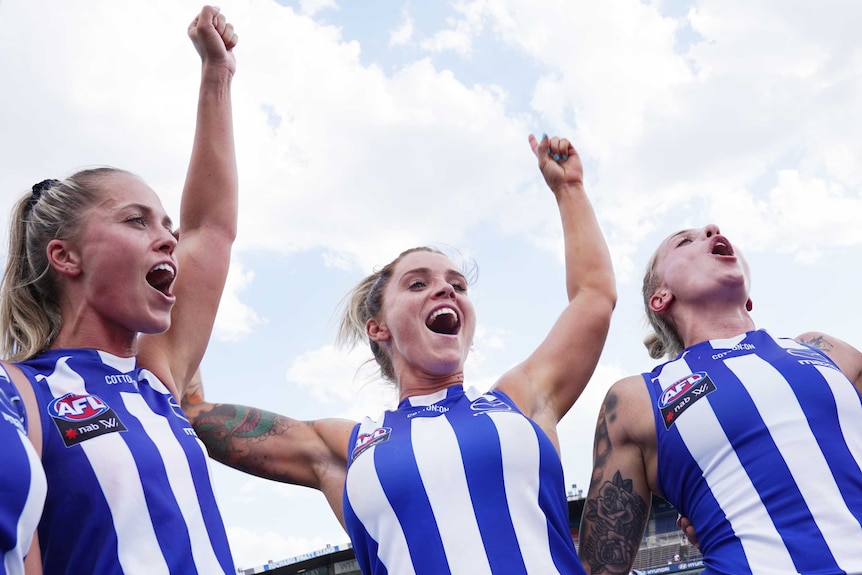 Sophie Abbatangelo, centre, holds both her arms up in the air wearing a blue and while singlet with two players next to her