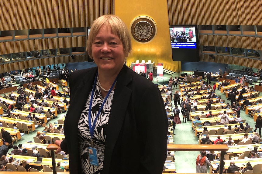 A woman with short blonde hair smiles for the camera with a parliament setting in the background.