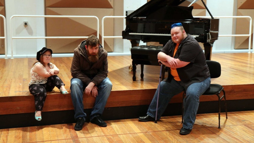 From left to right, Emma J Hawkins and Scott Price sit on a stage with a piano, Erin Kyan sits on a chair in front of the stage.