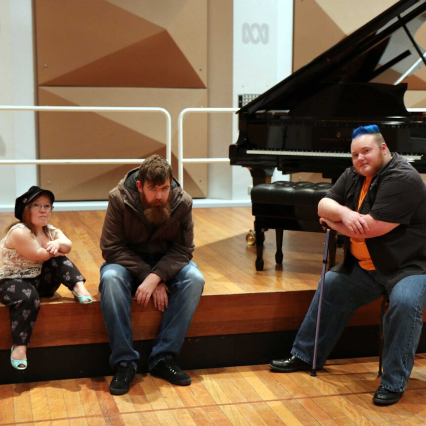 From left to right, Emma J Hawkins and Scott Price sit on a stage with a piano, Erin Kyan sits on a chair in front of the stage.