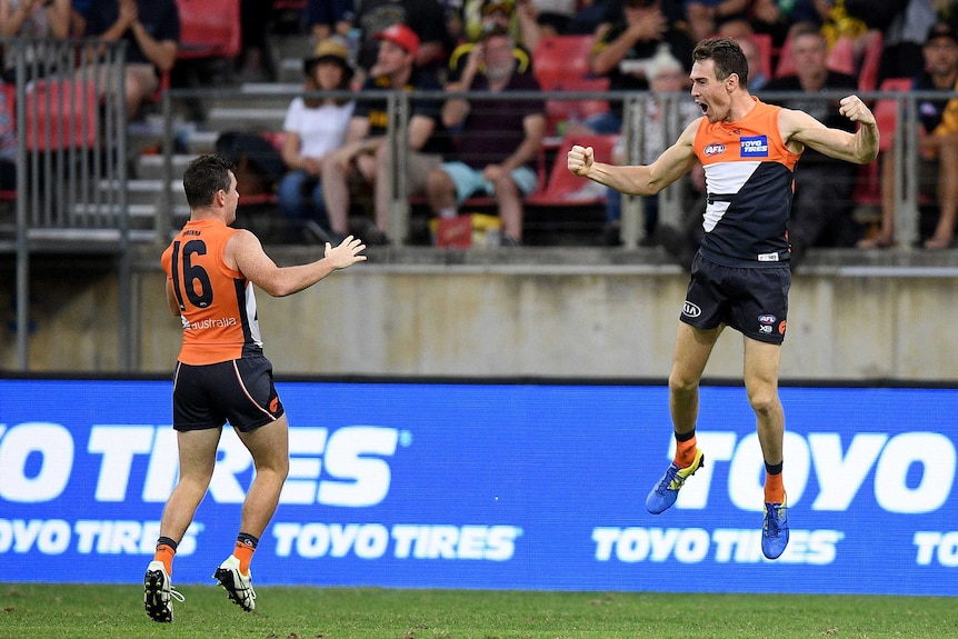 An AFL forward leaps in the air in delight after kicking a goal, as a teammate watches.