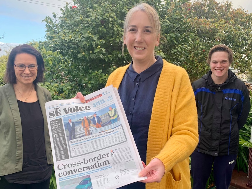 A woman stands presenting a newspaper, with two people standing proudly beside her.