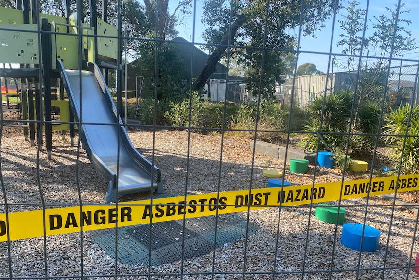 Yellow 'DANGER ASBESTOS DUST HAZARD' tape in front of a playground with a metal slide and gate.