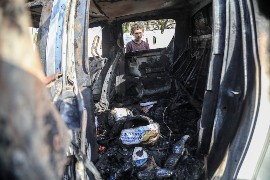 A young boy looks inside a destroyed car, seen through a window from the other side of the vehicle